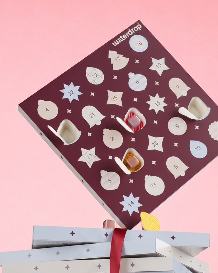 Purpurový Adventní kalendář se hvězdičkovými dveřmi na růžovém pozadí, nad dvěma zabalenými dárky.