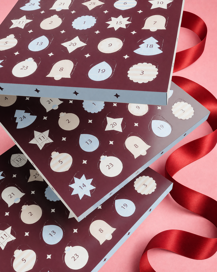 Tři na sebe položené kalendáře Advent s očíslovanými dvířky a dekorativními tvary, ozdobeny červenou stuhou na růžovém pozadí.
