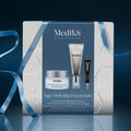 A Medik8 "Age-Defying Essentials" gift box wrapped with a blue ribbon on a reflective surface against a dark blue background. The white box features a dark blue center panel displaying the printed products inside: an Advanced Pro-Collagen+ Peptide Cream jar, a Crystal Retinal 6 tube, and a Liquid Peptides Advanced MP deluxe sample. Text highlights "Ultimate Skin Renewing Heroes."