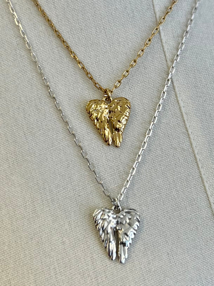 Two angel wing pendants on gold and silver necklaces against a light background