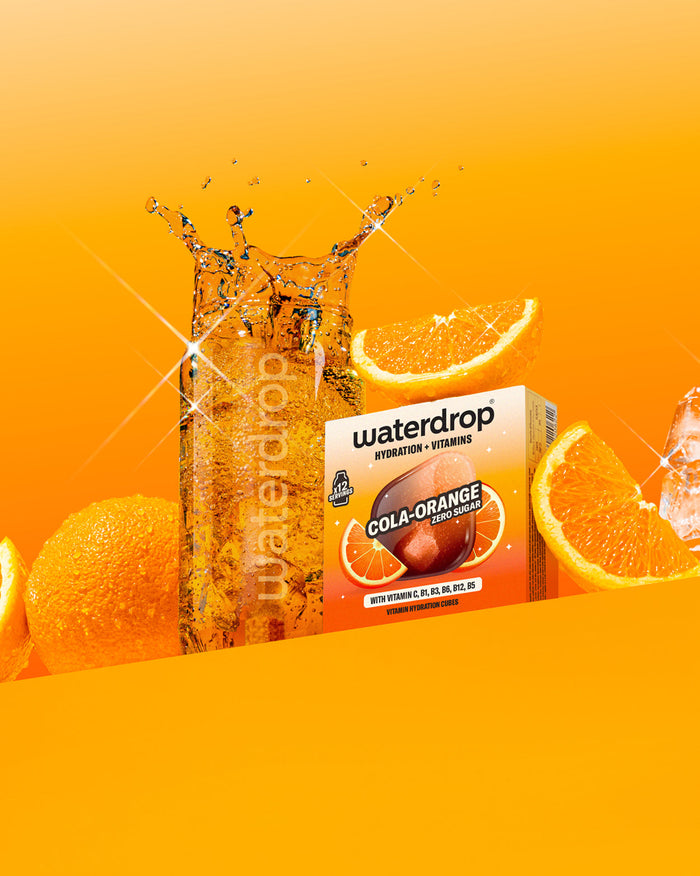 Hydratační kostky COLA ORANGE s vitamíny vedle perlivé vody a pomerančů.