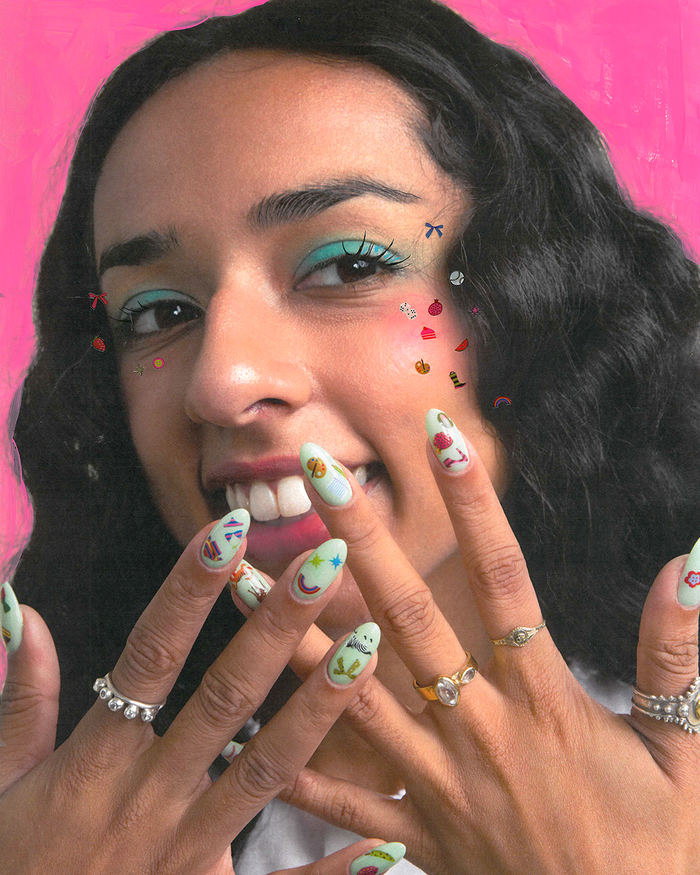 A close-up lifestyle shot of a woman with a bright smile, showing her nails with nail art stickers, along with silver rings, set against a vibrant pink background.