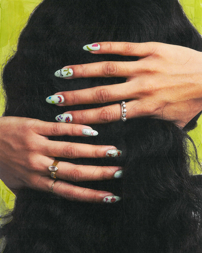 Close-up of a person's hands with long black hair, decorated with nail art stickers and small rings, against a bright yellow-green background.