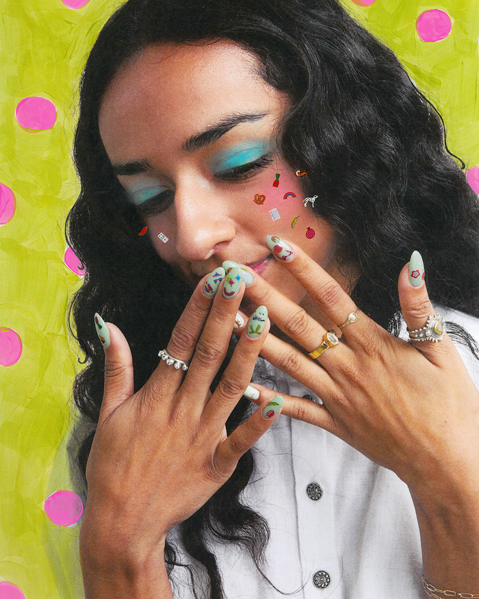 A close-up portrait of a woman with dark wavy hair, turquoise eye makeup, and stickered nails, wearing multiple rings, set against a bright lime green background with pink polka dots.