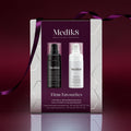 A Medik8 "Firm Favourites" gift box wrapped with a purple ribbon, set against a deep burgundy background with a reflective surface. The white box features a burgundy center panel displaying a Liquid Peptides Advanced MP black pump bottle and an Exo-PDRN Prismatic+ white pump bottle inside.