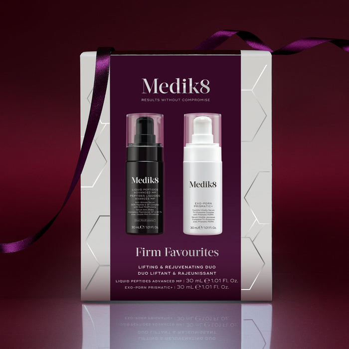 A Medik8 "Firm Favourites" gift box wrapped with a purple ribbon, set against a deep burgundy background with a reflective surface. The white box features a burgundy center panel displaying a Liquid Peptides Advanced MP black pump bottle and an Exo-PDRN Prismatic+ white pump bottle inside.