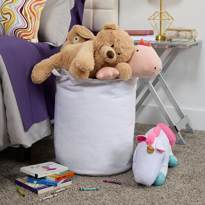 White laundry hamper filled with stuffed animals on a carpeted floor next to a bed.