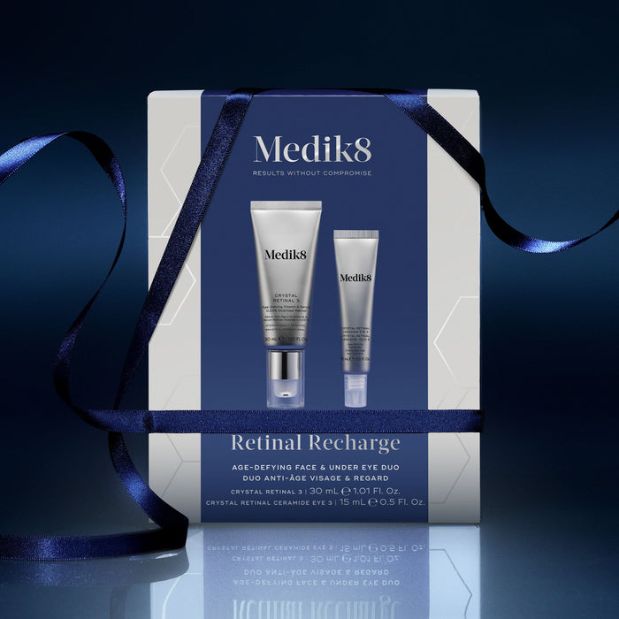 A Medik8 "Retinal Recharge" gift box wrapped with a dark blue ribbon on a reflective surface against a dark blue background. The white box features a dark blue center panel displaying the printed products inside: a Crystal Retinal 3 tube and a Crystal Retinal Ceramide Eye 3 tube. Text highlights "Age-Defying Face & Under Eye Duo."