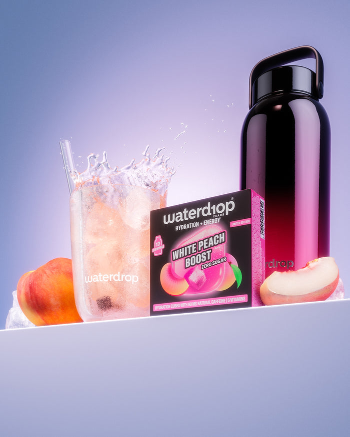 Balíček Waterdrop s peachovou příchutí vedle sklenice s výměšky, tmavé lahve a plátků broskve.