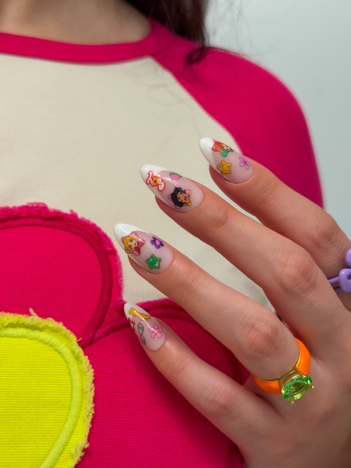 Close-up of a hand with colorful nail art stickers wearing a bright orange ring, against a pink and yellow background.