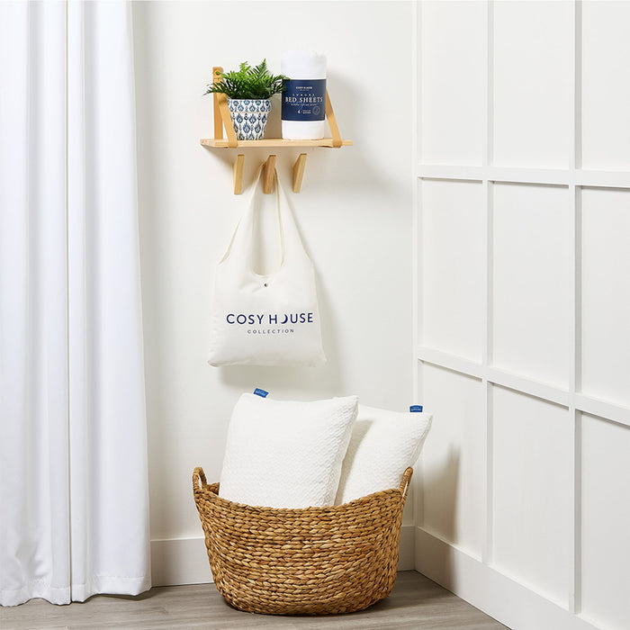 Basket with pillows, plant, and 'Cosy House' tote bag against a white wall.